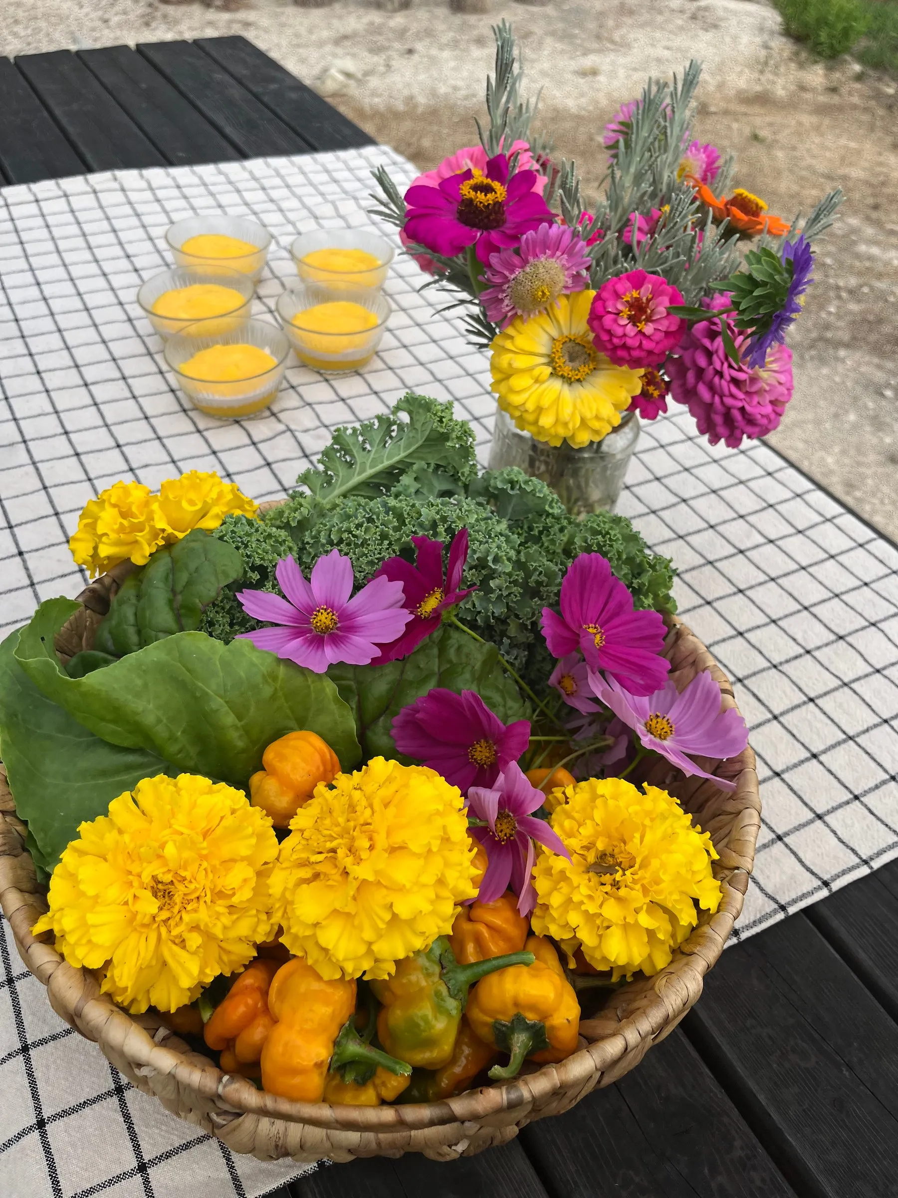 A basket of marigolds, cosmos, kale and small peppers on the table, with a jar of zinnias and desserts behind it